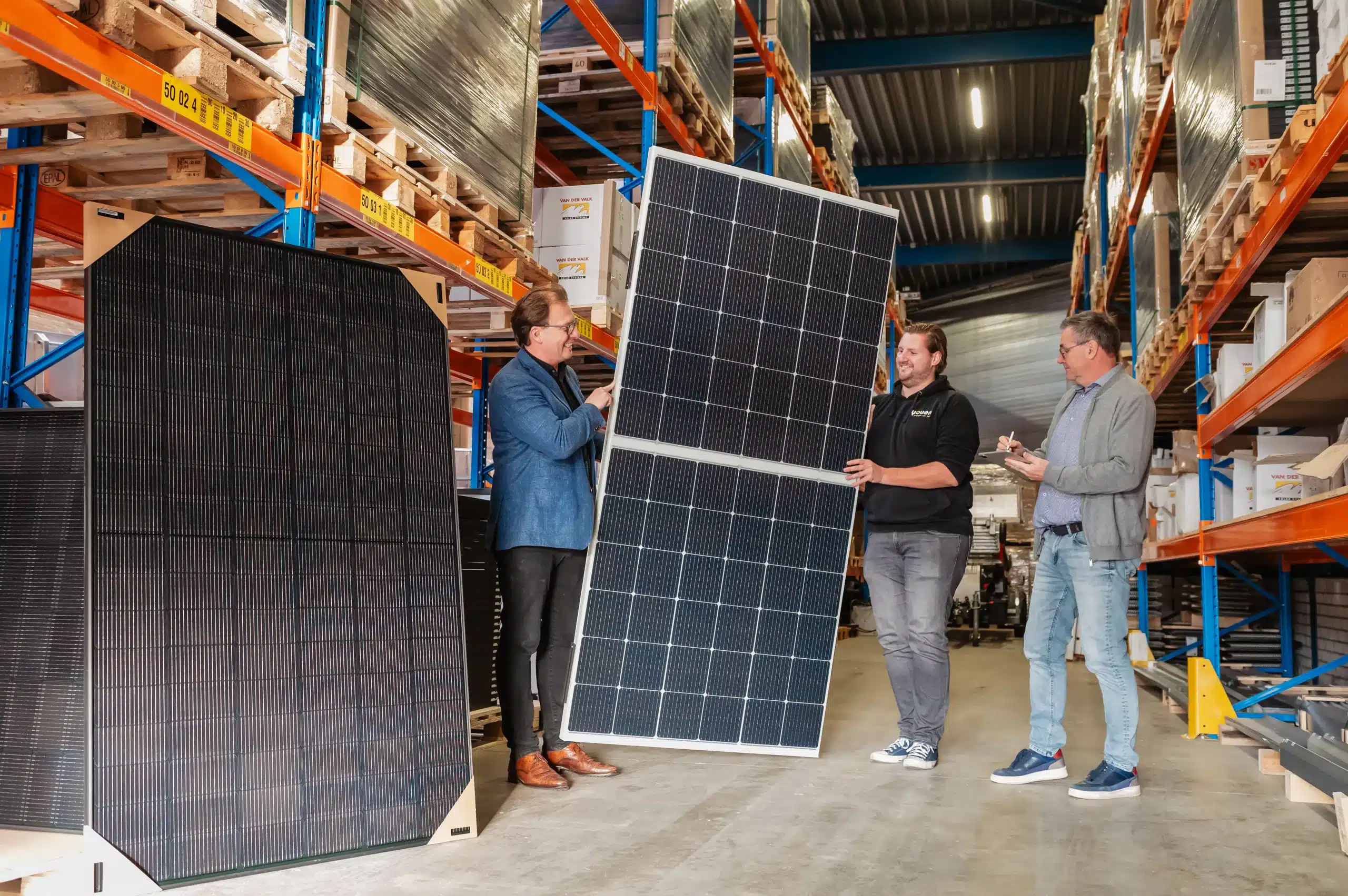 Drie professionals bespreken zonnepanelen in een magazijn met op de achtergrond schappen gevuld met zonnepanelen.
